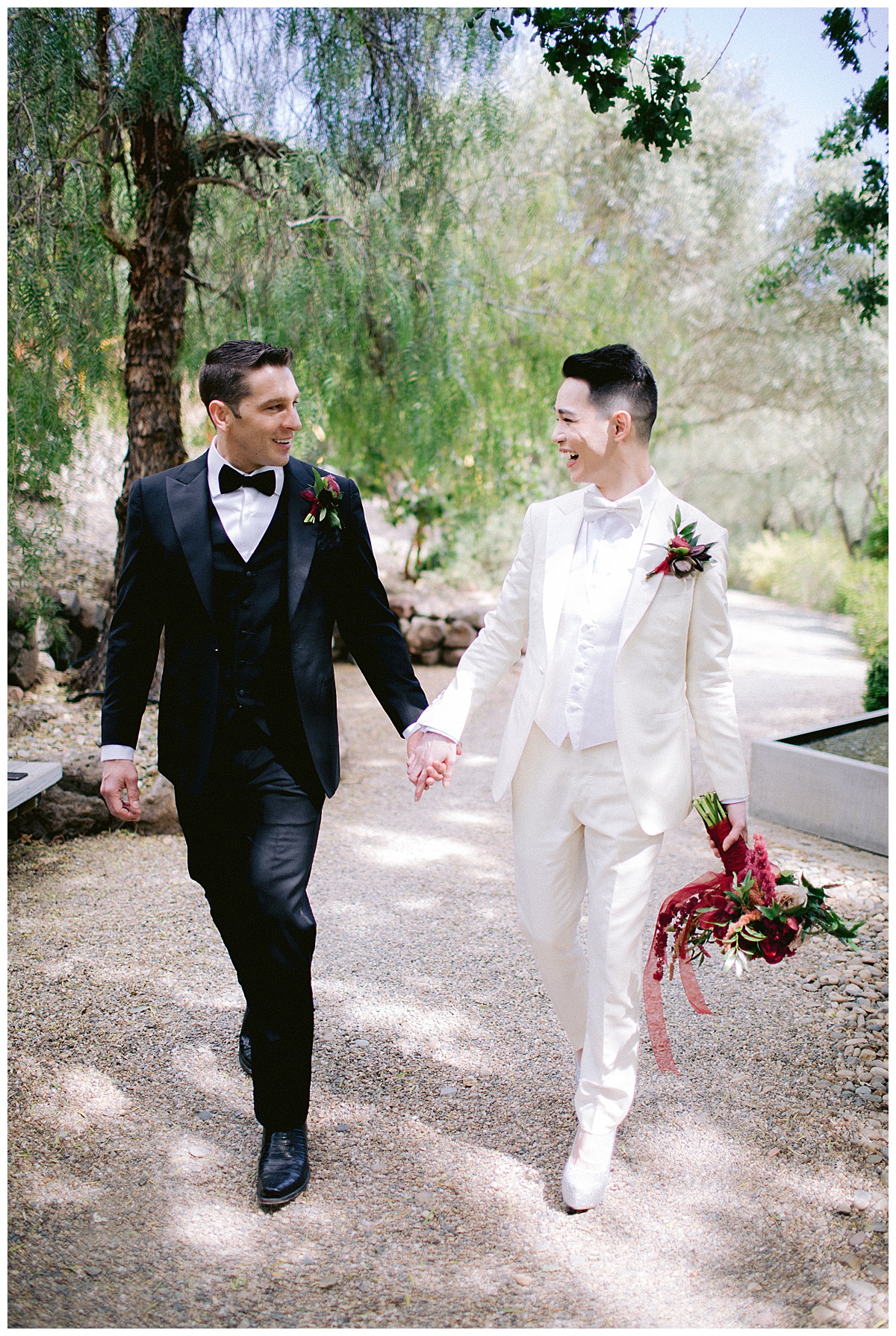 Groom in black and and groom with burgundy bouquet hold hands smiling in a park in the San Francisco Bay Area