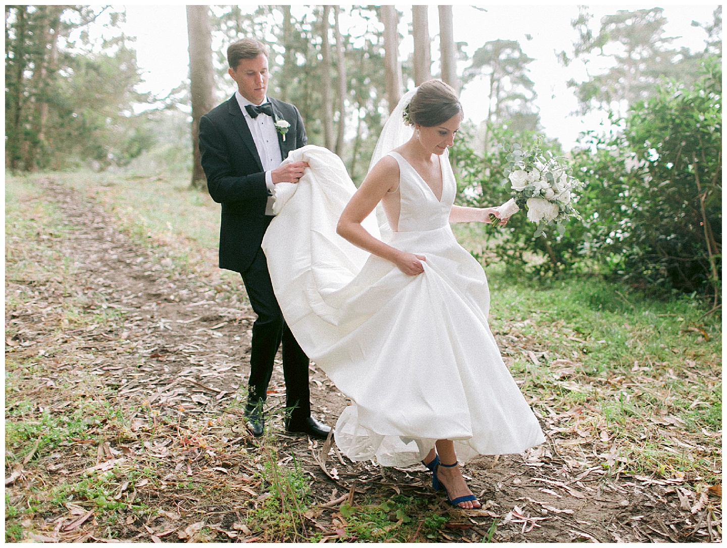 groom holds brides dress while walking through the woods